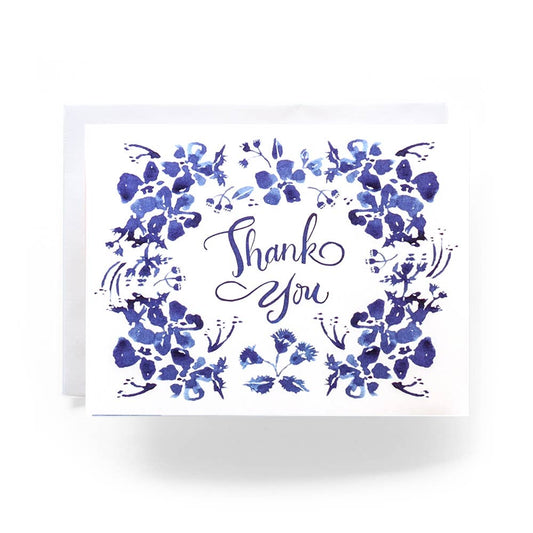 A thank you card by Antiquaria. The base of the card is a soft white with blue wildflowers around the border of the card. Thank you is written in blue in the center.